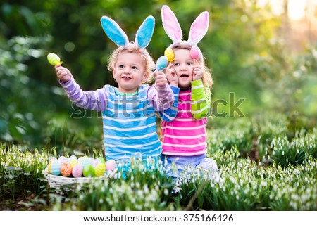 Kids on Easter egg hunt in blooming spring garden. Children with bunny ears searching for colorful eggs in snow drop flower meadow. Toddler boy and preschooler girl in rabbit costume play outdoors.  - stock photo