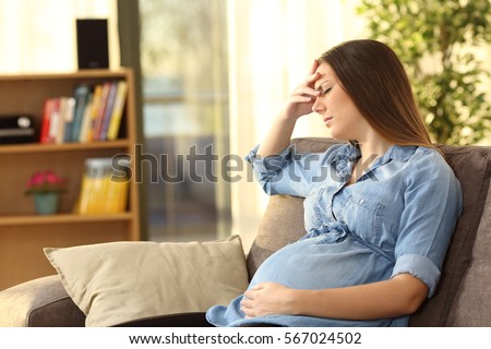 Pregnant woman with hand on forehead suffering headache sitting on a couch in the living room at home