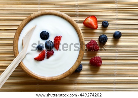 Wooden bowl of white yogurt with wooden spoon inside on bamboo matt from above. Next to berries.