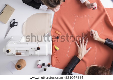 Top view of two girls making a pattern on a red piece of material. Sewing machine is standing on the table beside them.