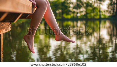 Woman relaxes by the lake sitting on the edge of a wooden jetty , swing one's feet near the water surface. Sunny joyful summer day or evening concept.