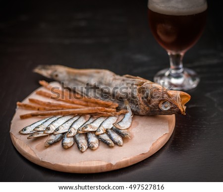 Glass of beer and assorted dried fish on a cooking sheet, traditional beer snack. Black wooden background.