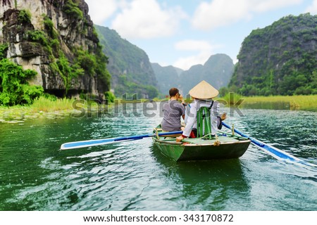 Tourists traveling in small boat along the Ngo Dong River at the Tam Coc portion, Ninh Binh Province, Vietnam. Rower using her feet to propel oars. Landscape formed by karst towers and rice fields.