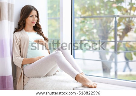 Young beautiful pregnant woman sitting on windowsill at home