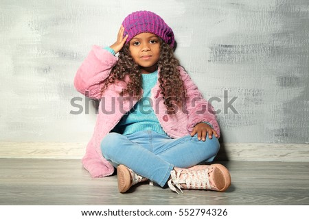 Cute little African American girl sitting against grey wall. Fashion concept