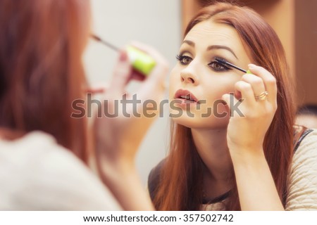 Brunette woman applying make up (paint her eyelashes) for a evening date in front of a mirror. Focus on her reflection