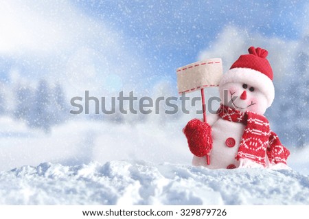 Winter arrival concept with Snowman with a blank placard in his hand. Dressed with red scarf and gloves and hat in the snow outside under the snow. Front view. Horizontal composition