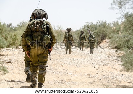 Israeli army combat soldiers return to base after completing military mission during military campaign / war. Desert landscape, armed infantry soldier walking in the foreground. Soldiers walking away.