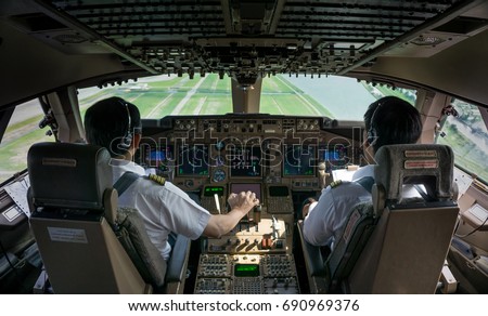 2 Asian Commercial pilots,captain and first officer (co pilot) fly the jet plane in daytime view in cockpit (flight deck) with green field outside as background.