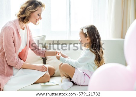 Cute little girl putting jewelry on hand of happy mother while sitting on sofa