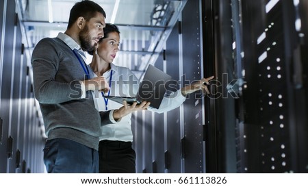 Male IT Specialist Holds Laptop and Discusses Work with Female Server Technician. They're Standing in Data Center, Rack Server Cabinet is Open.