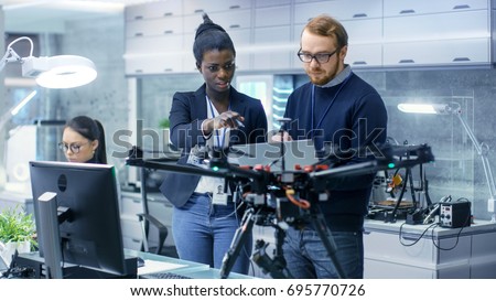 Caucasian Male and Black Female Engineers Working on a Drone Project with Help of Laptop and Taking Notes. He Works in a Bright Modern High-Tech Laboratory.