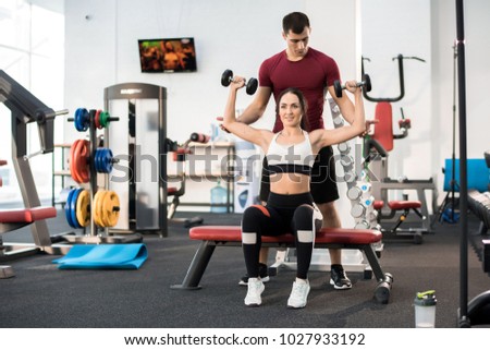 Full Length Portrait Of Beautiful Young Woman Exercising With Dumbbells Sitting On Bench Helped By Personal Full Length Portrait Of Beautiful Young Woman Exercising With Dumbbells Sitting On Bench Helped By Personal