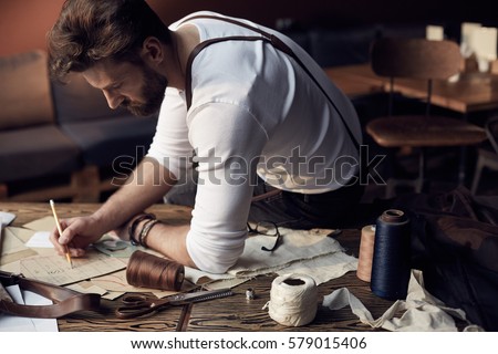 Handsome tailor male with beard in white shirt with brown leather suspenders working near wooden table with threads in amazing atelier with antique furniture and mannequin on background