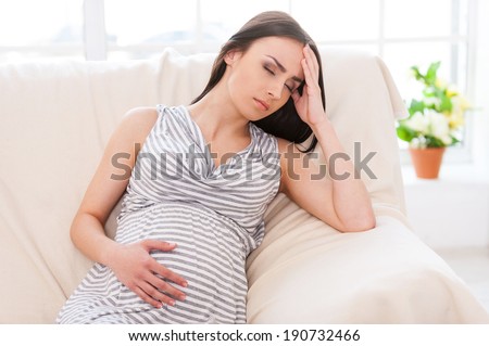 Struggling with morning sickness. Depressed pregnant woman holding hand on head and keeping eyes closed while sitting on a couch