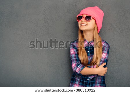 Stylish and cute. Cheerful little girl in sunglasses keeping arms crossed and looking up with smile while standing against grey background