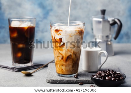 Ice coffee in a tall glass with cream poured over and coffee beans on a grey stone background