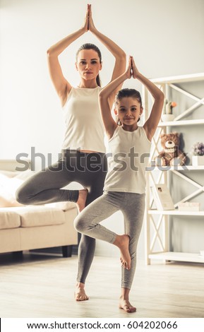 Beautiful young woman and charming little daughter are smiling while doing yoga together at home