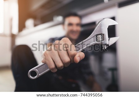 The plumber sits next to the kitchen sink on the floor and selects the appropriate spanner for repairing the pipe. He shows the wrench in the camera. The man has a special working uniform.