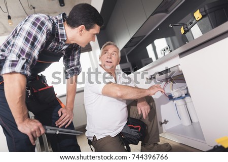 Two plumbers prepared to repair the kitchen sink. One of them is next to the pipes, the second is near and helps to choose a tool. A black toolbox is next to it.