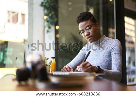 Concentrated afro american experienced woman journalist in glasses creating article working in coffee shop, skilled young female graphic designer making sketch in notebook for new startup project