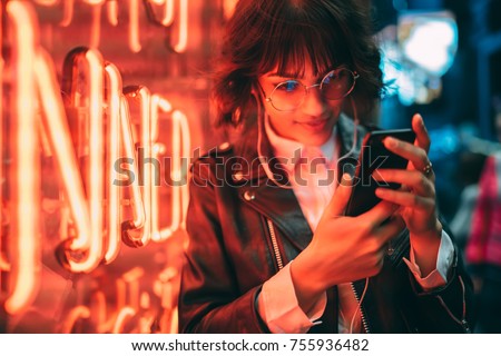 Young stylish female hipster standing near neon red light in night city and listening to music in headphones on modern smartphone. Woman watching media video on cellphone device