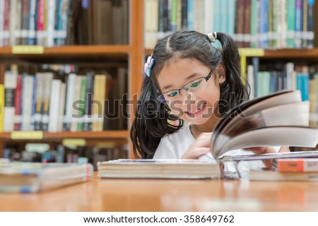 Happy little asian child girl wear eyeglasses reading book school background: Lovely cute young student kid opening flipping book in archive resource collection room: National library lover week