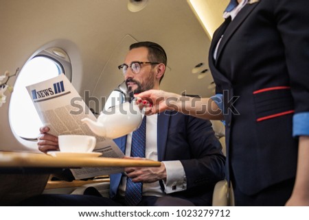 Glad groomed businessman reading newspaper in airplane while stewardess is serving tea