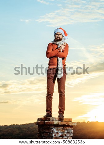 young bearded santa claus man with long beard in red sweater pants and new year hat in scarf with glass of champagne drinking and celebrating christmas or xmas on sunny blue sky on brick chimney