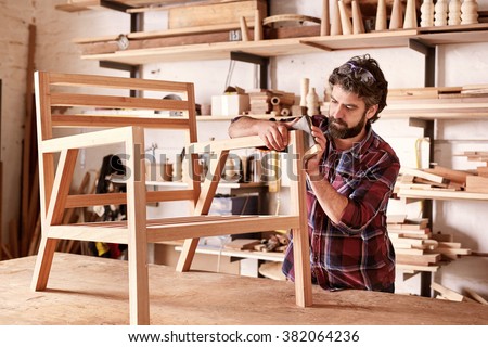 Serious furniture designer carefully sanding a chair frame that he is busy manufacturing in his woodwork studio, with shelves of wooden items behind him