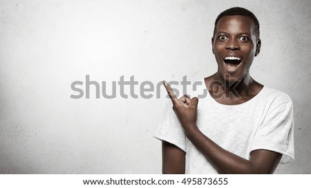 Headshot of attractive African man looking at camera with amazed expression and full disbelief, pointing his index finger at blank copy space wall, shocked with content on it, saying: Look at that!