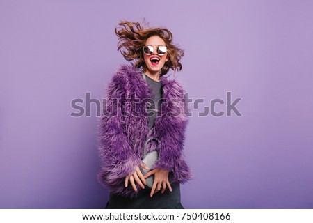 Gorgeous short-haired girl in sunglasses dancing on purple background with happy smile. Laughing female model in elegant fur coat posing in studio with little handbag.