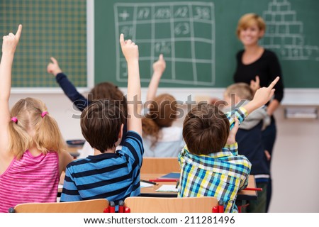 Intelligent group of young school children all raising their hands in the air to answer a question posed by the female teacher, view from behind