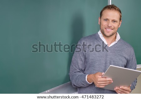 Happy friendly young teacher or pst graduate male student standing holding a tablet in front of a blank chalkboard