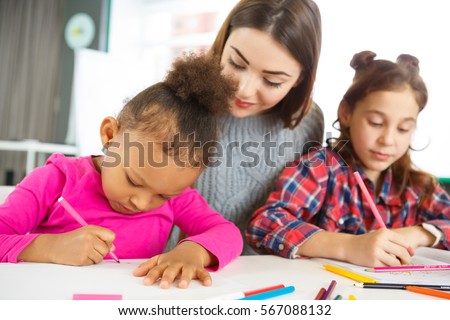 Teaching kids. Attractive cheerful young female teacher smiling watching her young student doing assignment on the lesson school classroom teaching learning education happiness childhood concept
