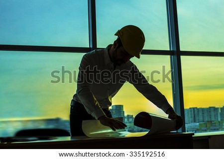 Silhouette builder engineer wear security helmet look at blueprint paper construction drawing plan on background of sunset window frame blue yellow sky with clouds near pc monitor computer
