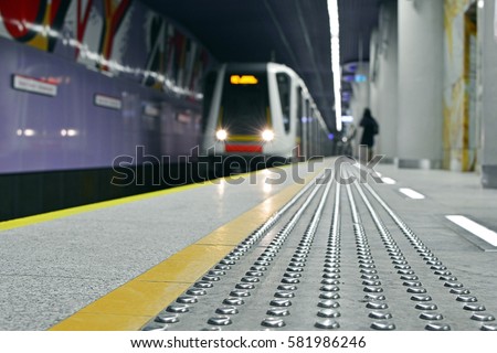 Metal tactile strips with an yellow line - for visually impaired (handicapped) and people with blindness - on an underground station, with a blurred arriving subway train and background - Warsaw Metro