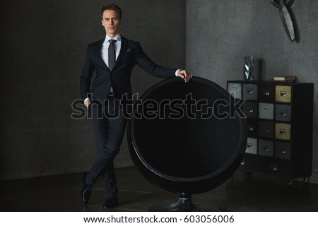 young businessman in black suit posing near the chair on gray background