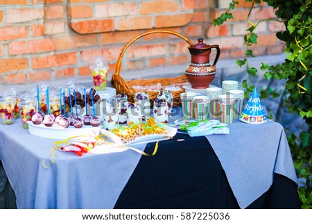 Photo decorated table for children's parties, birthday against backdrop of orange brick wall. In photo depicts variety treats, delicious food, clear jars with cottage cheese soufflé with layer of