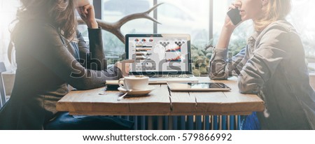 Teamwork, two young businesswomen sitting at table. On table laptop, coffee cup and tablet computer. First girl showing pencil on graphs and charts on computer screen, other is talking on cell phone.