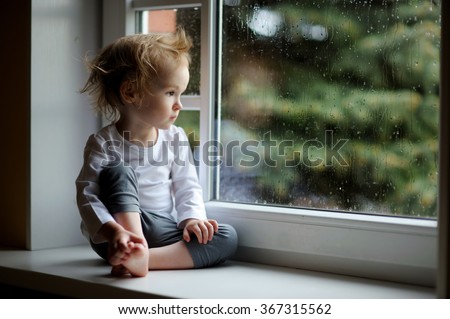 Adorable toddler girl looking at raindrops on the window