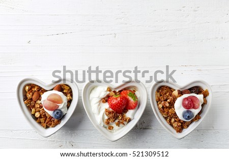 Heart shaped bowls of homemade granola and yogurt on white wooden background from top view