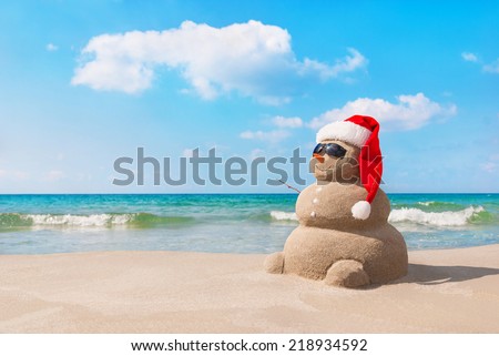 Sandy christmas snowman in red santa hat and sunglasses at sunny beach. Holiday concept for New Years Cards.