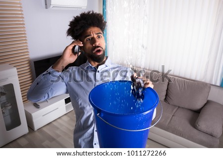 Worried Young Man Calling Plumber While Leakage Water Falling Into Bucket At Home