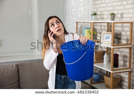 Worried Woman Calling Plumber While Collecting Water Droplets Leaking From Ceiling At Home