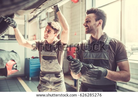 At the auto service. Handsome young auto mechanics in uniform are smiling while repairing car