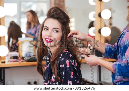 Female hairdresser standing and making hairstyle to cute lovely young woman in beauty salon