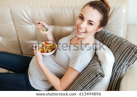 Happy pregnant young woman sitting and eating fruit salad on sofa at home