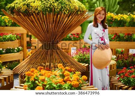 Culture Of Asia. Beautiful Happy Smiling Young Asian Woman Wearing Traditional White Ao Dai Dress ( Clothing ), Vietnamese Conical Hat ( Non La ) Leaf Hat In Flower Garden In Ho Chi Minh City, Vietnam