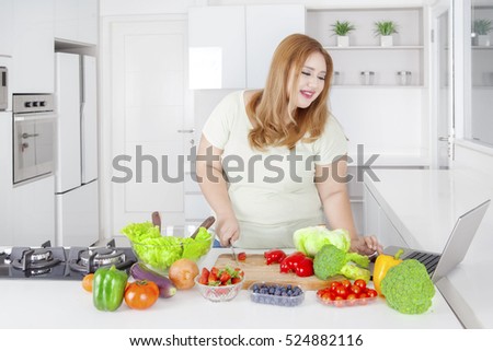 Portrait of obese woman cutting paprika by using knife while looking receipt at laptop 